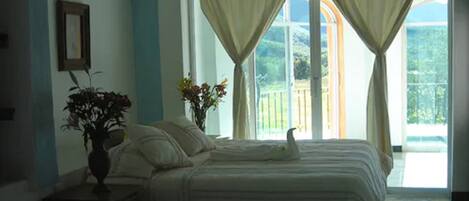 Quarto com cama, mesa de cabeceira, vaso com flores e janela com cortinas.