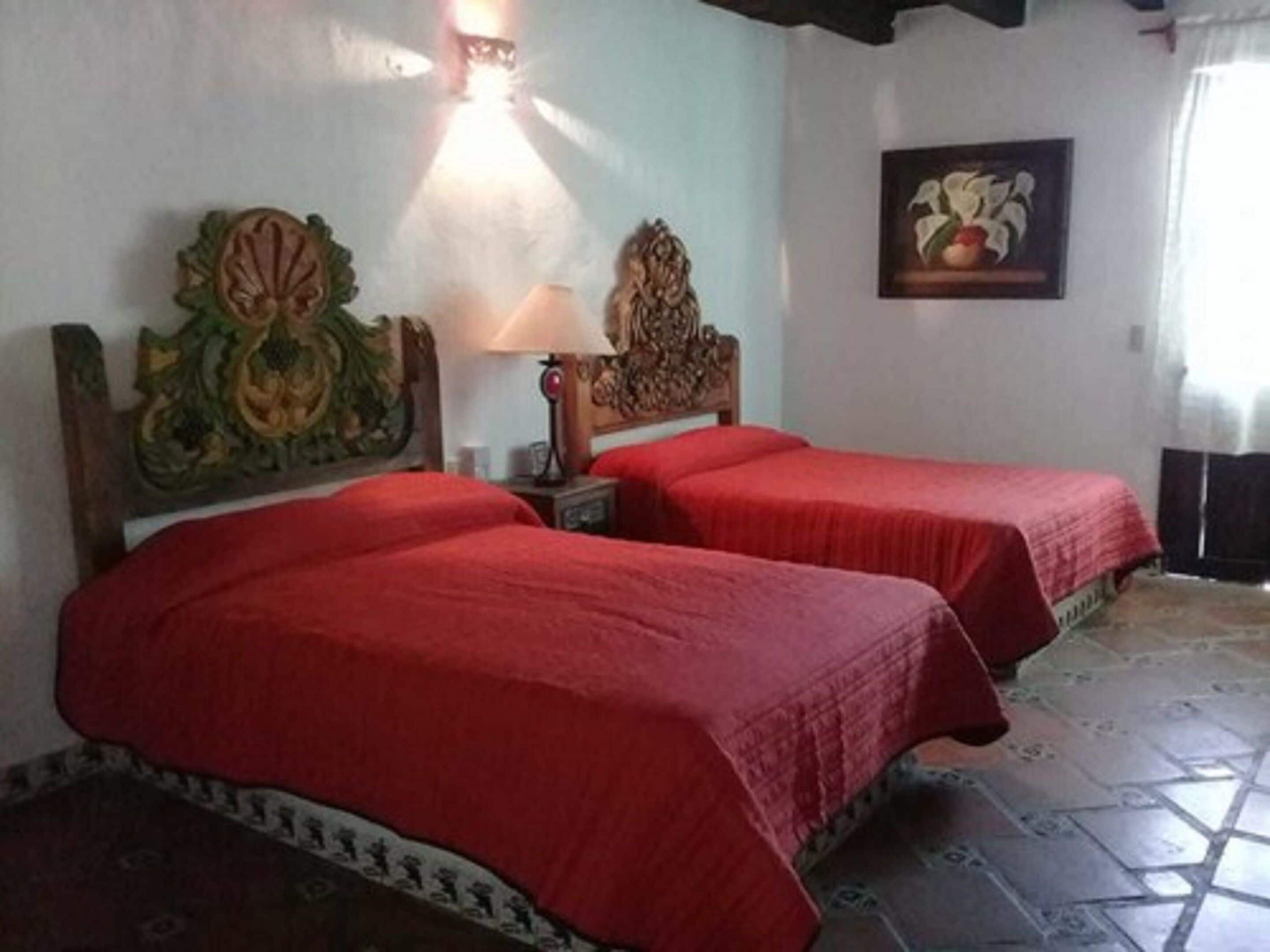 Ein Zimmer mit zwei Betten, jeweils mit roter Bettdecke, einem hölzernen Kopfstück mit aufwendigen Schnitzereien, einem Nachttisch mit Lampe und einem gerahmten Bild an der Wand.