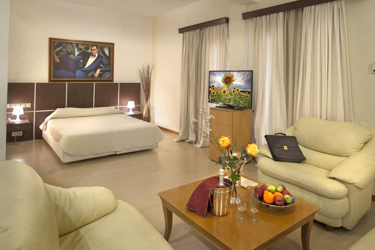A hotel room with a bed, a sofa, a coffee table with a vase and fruit bowl, a television, and a painting on the wall.