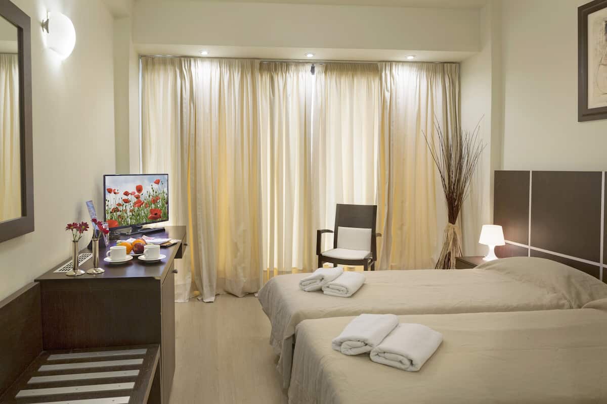 A hotel room with two beds, a desk with a computer and a vase of flowers, a chair, and a large window with curtains.