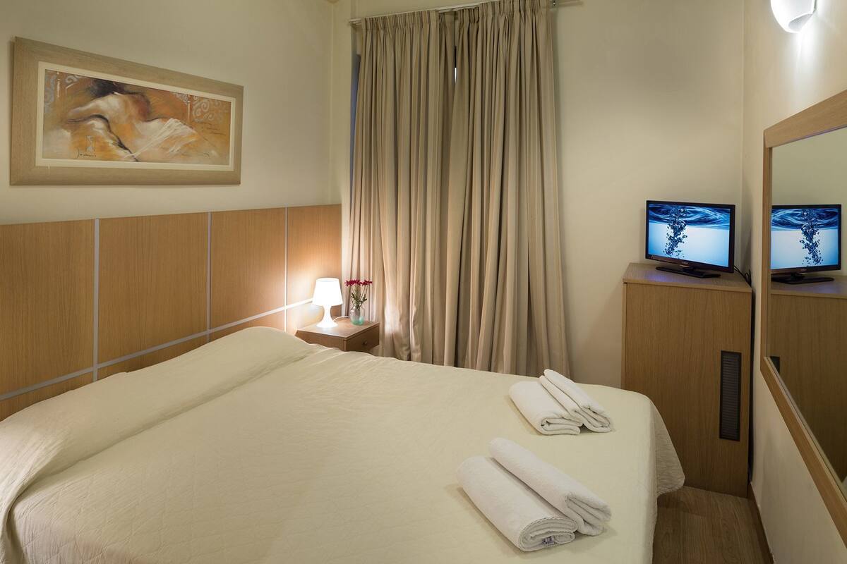 A hotel room with a bed, a nightstand with a lamp and flowers, a TV on a wooden cabinet, and a painting on the wall.