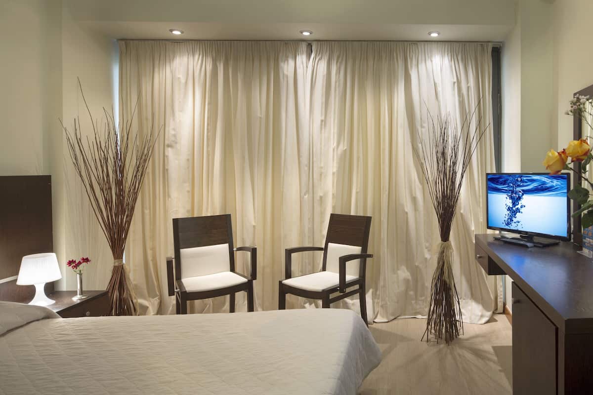 A hotel room with a bed, two chairs, a desk with a computer, a lamp, and a vase with flowers.