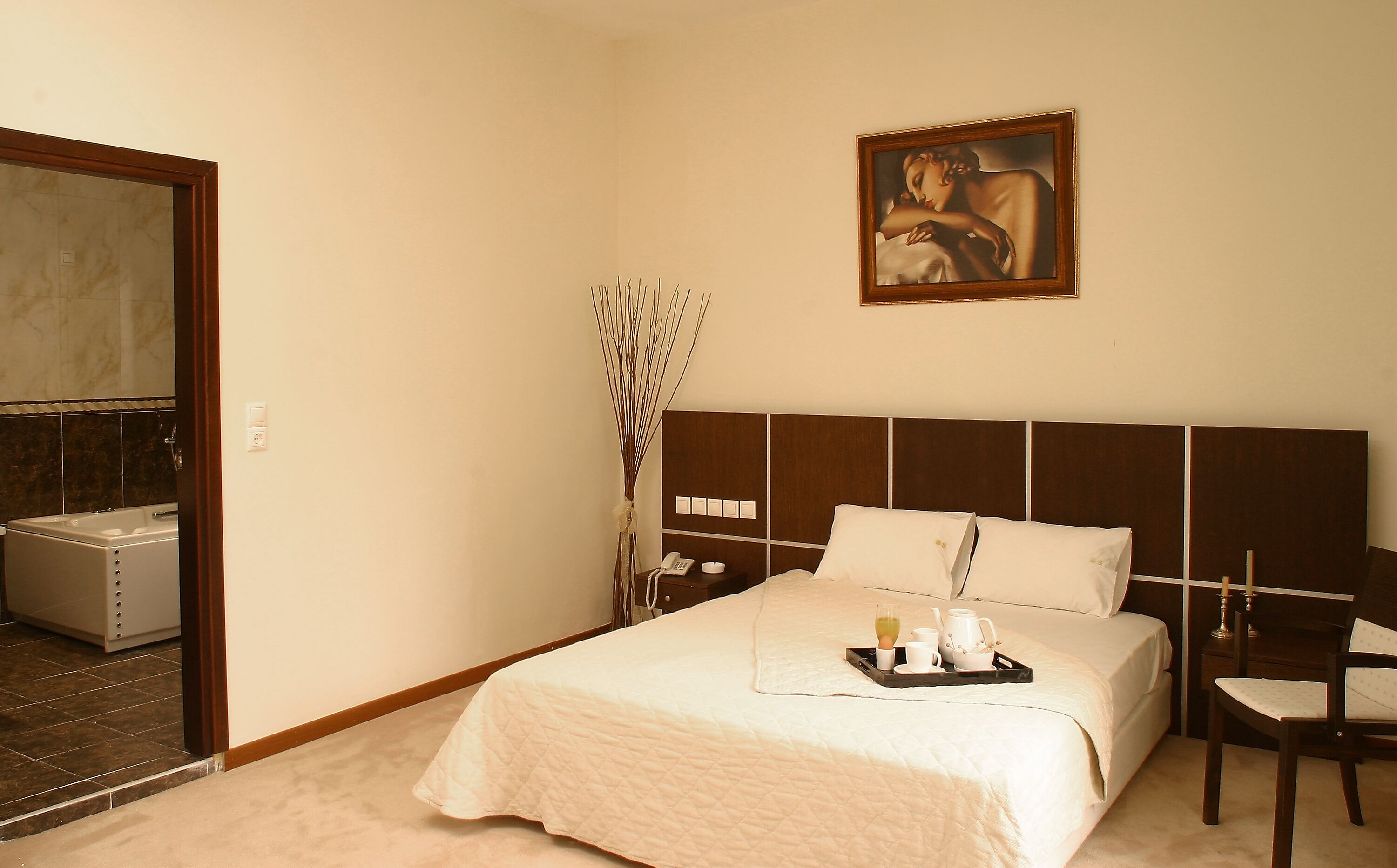 Una habitación de hotel con cama, mesita de noche, un cuadro y baño visible a través de una puerta abierta.
