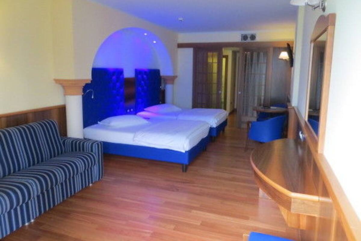 Quarto de hotel com piso de madeira, sofá, duas camas, um espelho e banheiro com chuveiro.