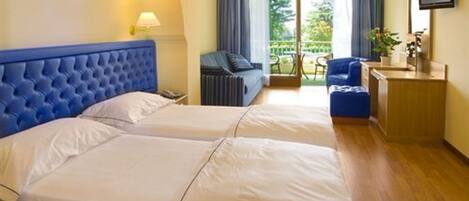 A hotel room with two beds, a desk, a chair, and a balcony with a view of greenery.