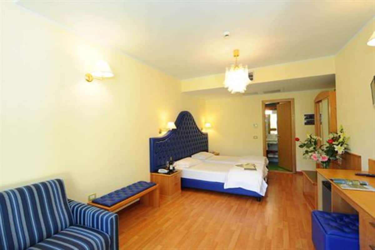 Quarto de hotel com uma cama de solteiro, mesa de cabeceira, cadeira, escrivaninha e uma pequena mesa com flores.