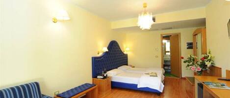 A hotel room with a single bed, a bedside table, a chair, a desk, and a small table with flowers.