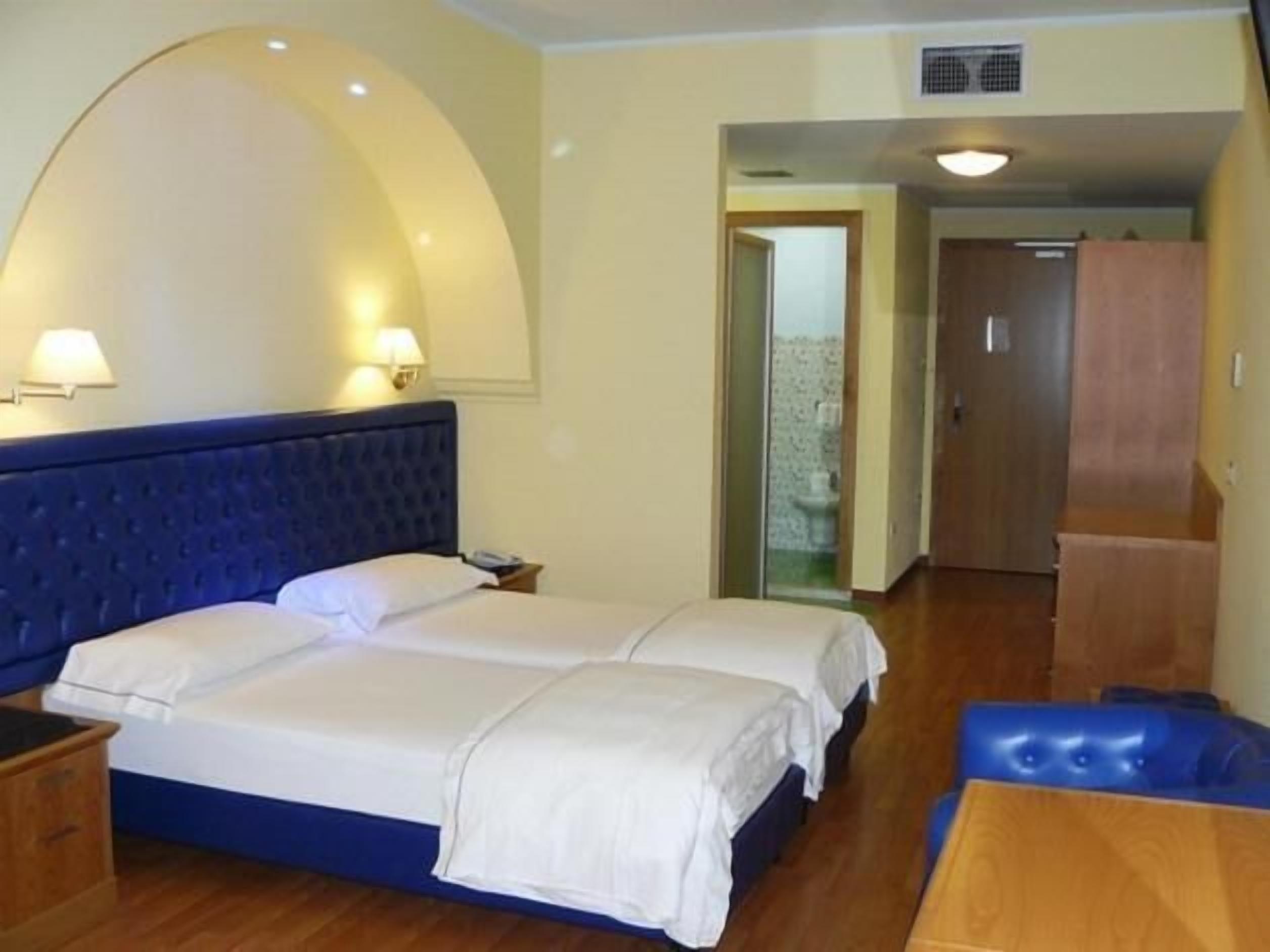 Una habitació d'hotel amb un llit gran, dues taules de nit, una panxa de fusta i un bany visible a través d'una porta oberta.