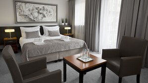 Een hotelkamer met een opgemaakt bed, twee nachtkastjes met lampen, een salontafel, twee fauteuils en een groot raam met gordijnen.