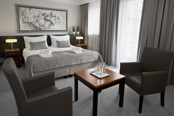 A hotel room with a neatly made bed, two bedside tables with lamps, a coffee table, two armchairs, and a large window with curtains.