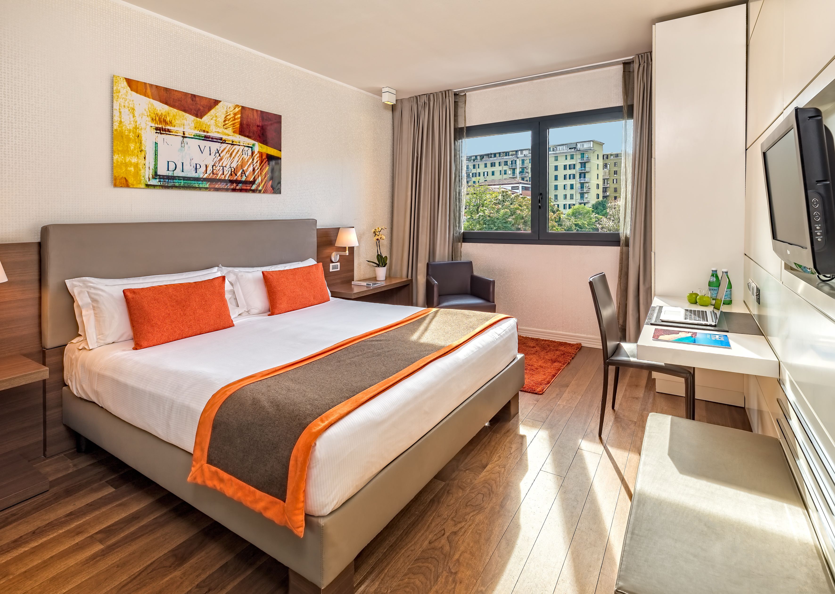 A hotel room with a large bed, a desk with a laptop, a TV, and a window with a city view.