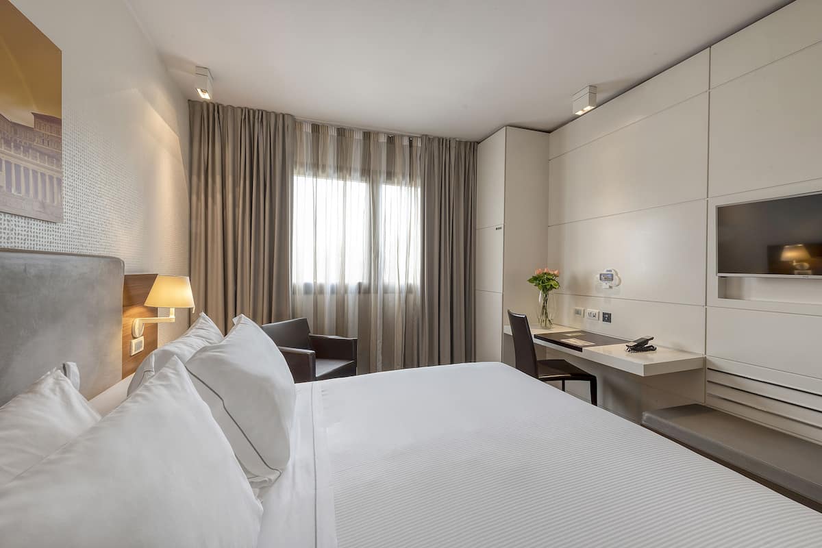 A hotel room with a large bed, a desk, a chair, a TV, and a window with curtains.