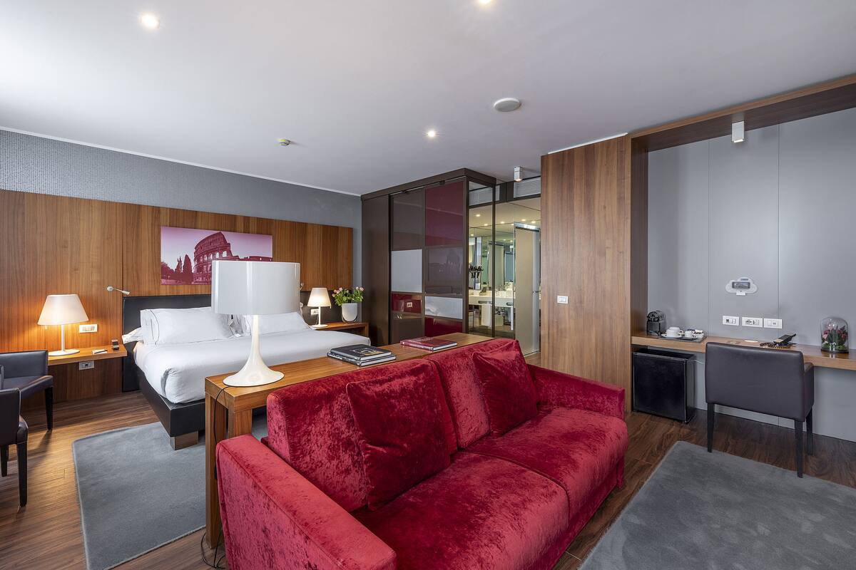A modern hotel room with a bed, a red velvet sofa, a desk with a lamp, and a small kitchenette.