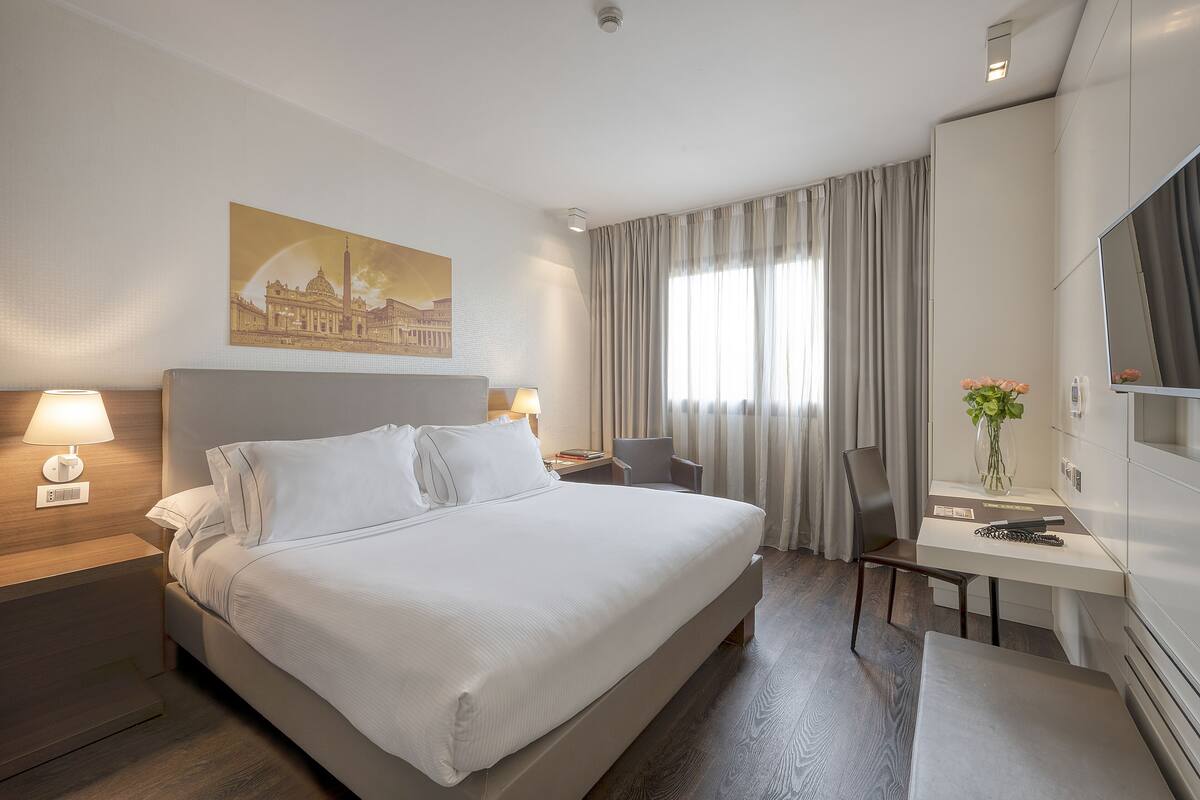 A hotel room with a large bed, bedside tables with lamps, a desk with a chair, a TV, and a painting on the wall.