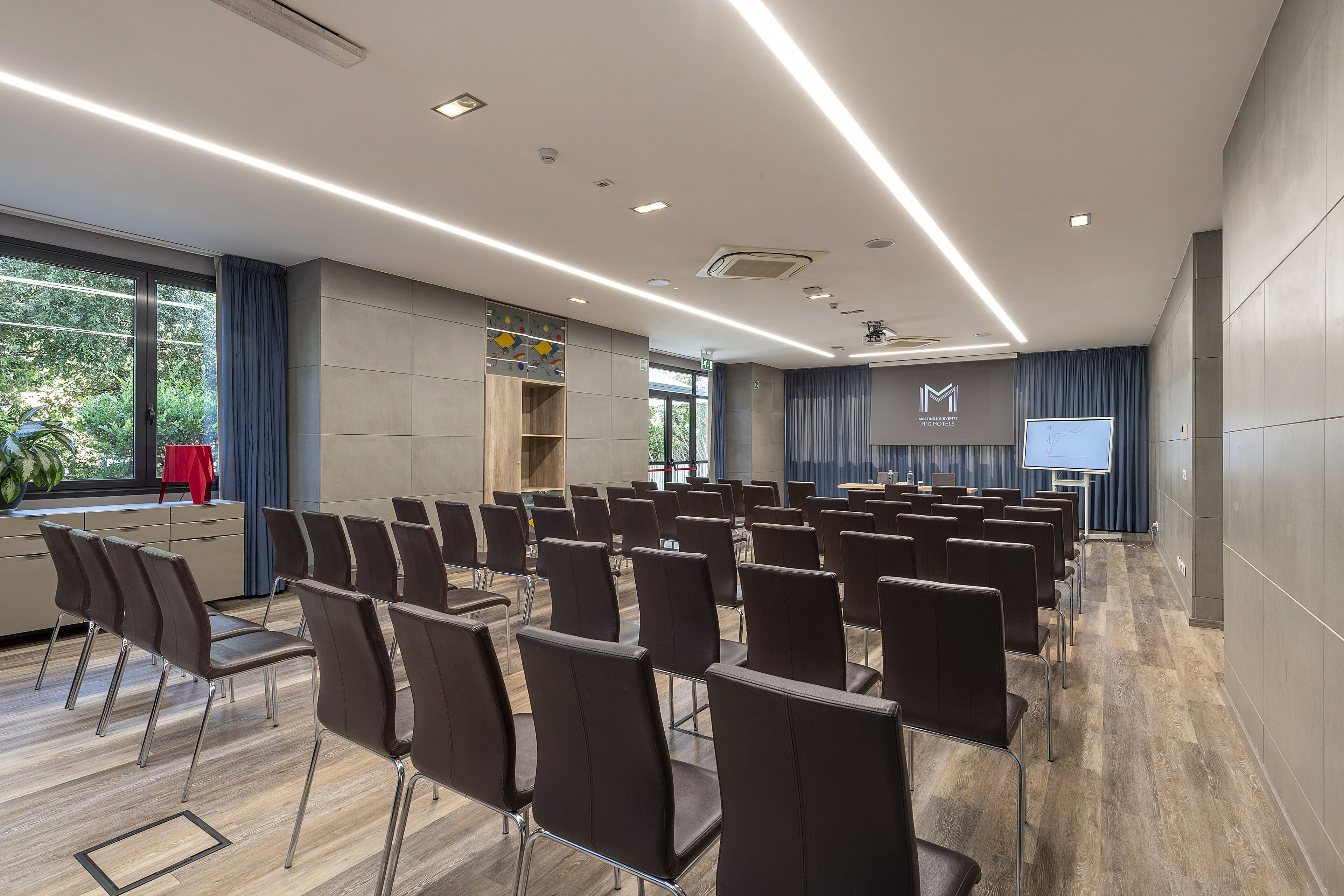 Un salón de conferencias con filas de sillas, una pantalla de proyector y un ventanal con cortinas.