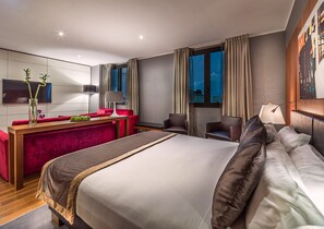 A hotel room with a large bed, a desk with a chair, a red sofa, and a TV mounted on the wall.