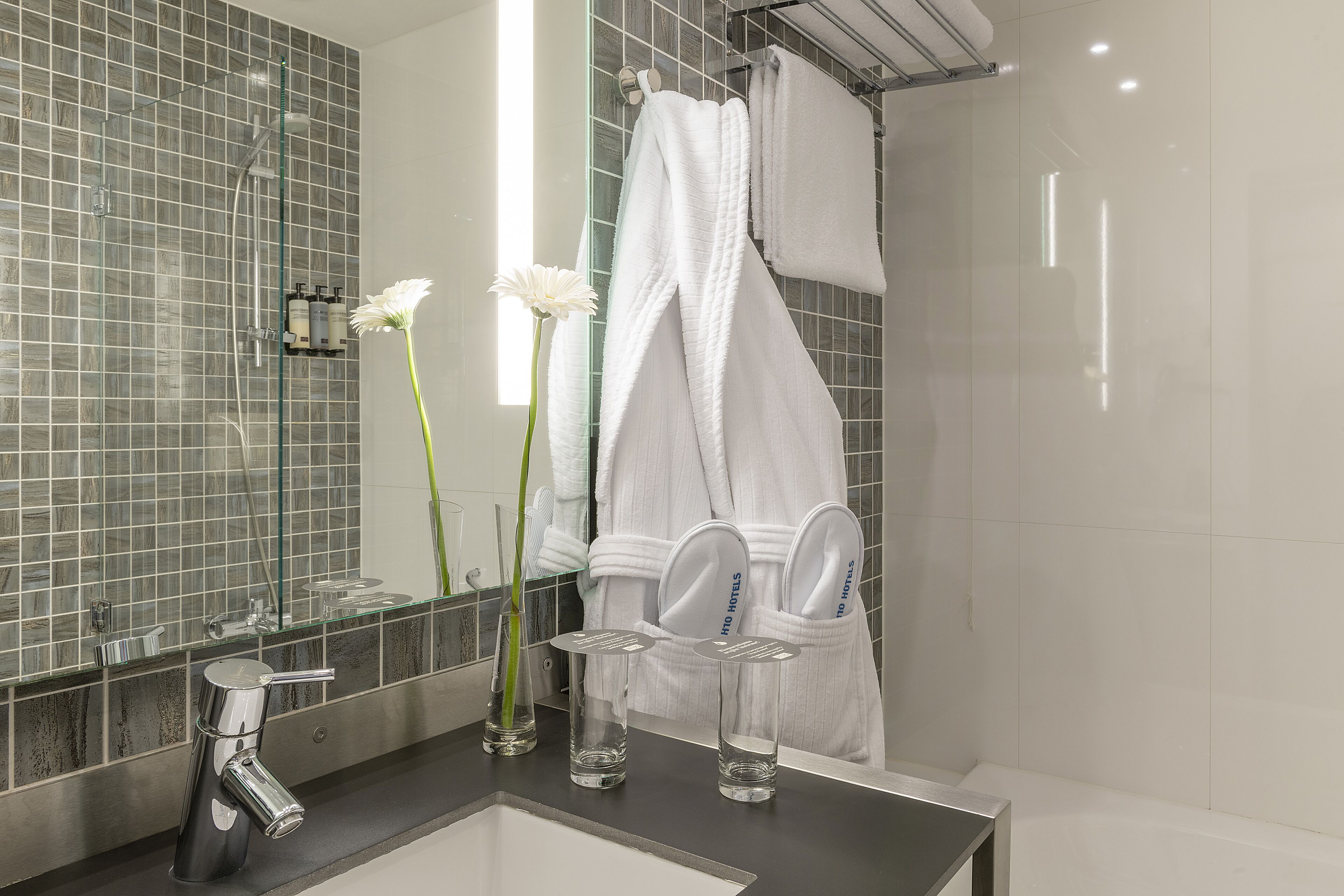 Un bagno moderno con cabina doccia in vetro, uno specchio grande e un mobile lavabo con lavandino e due bottiglie di vetro trasparente.