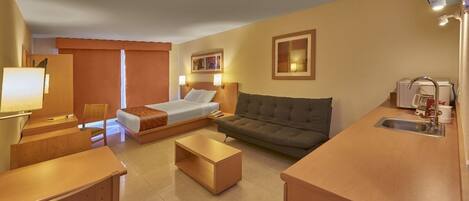 A compact hotel room with a bed, sofa, coffee table, and a kitchenette area.