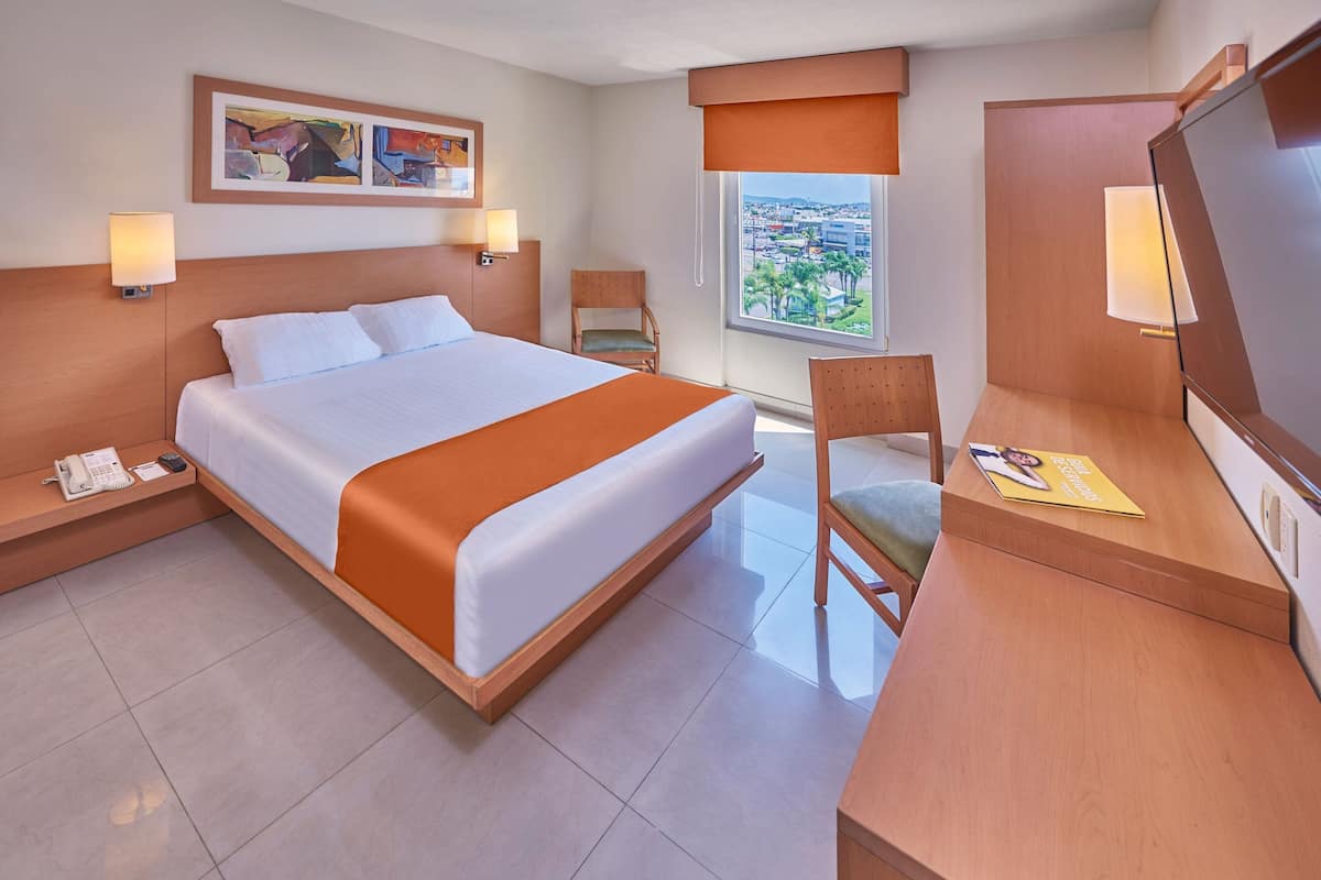 A hotel room with a large bed, a desk, a chair, a phone, and a window with a city view.