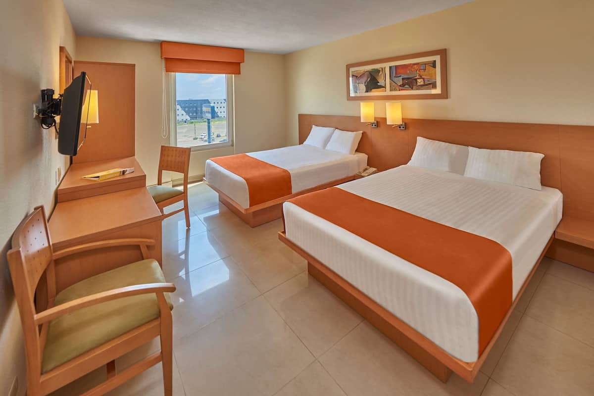 A hotel room with two beds, a desk, a chair, and a window with a view of buildings.