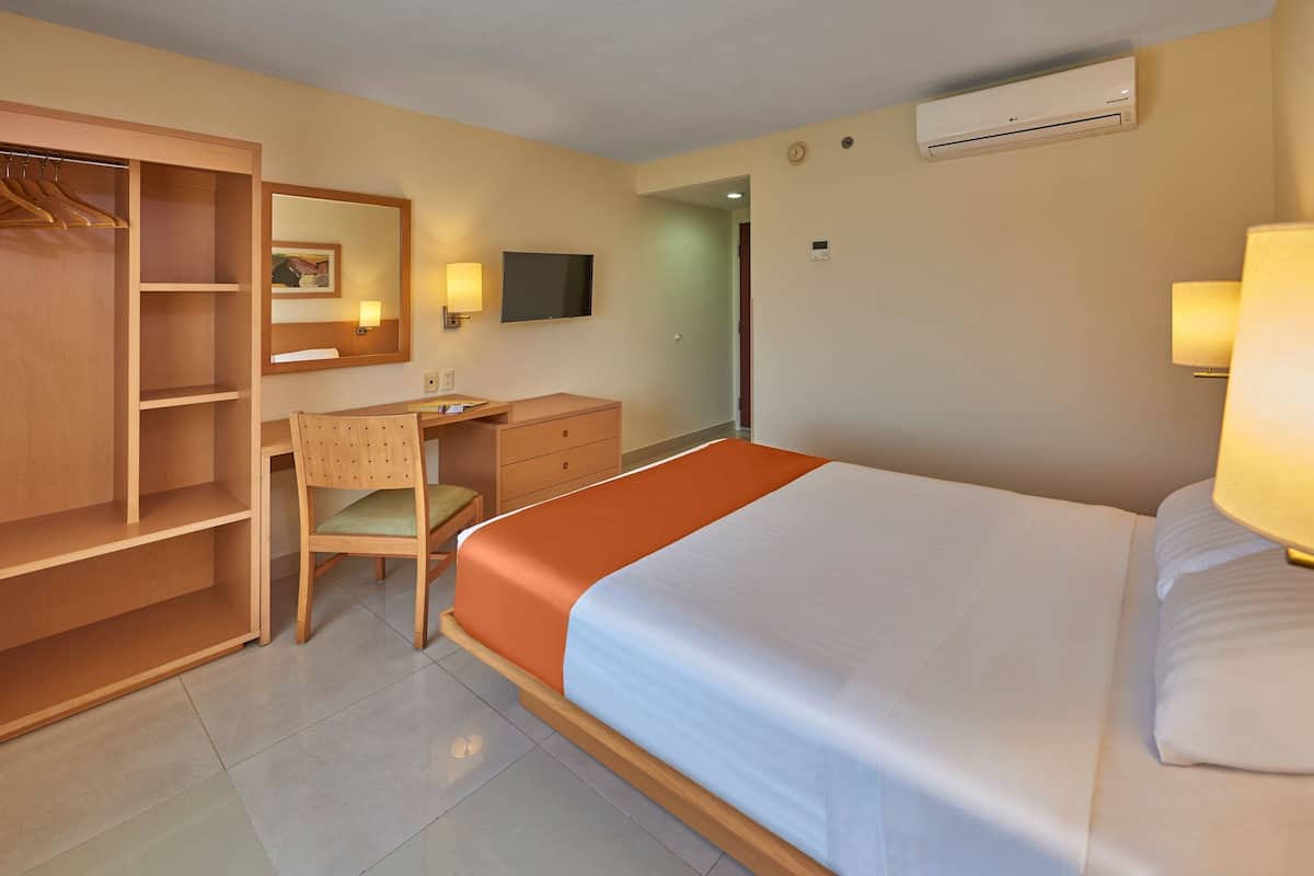 A hotel room with a bed, a desk, a chair, a wardrobe, a mirror, a television, and an air conditioning unit.