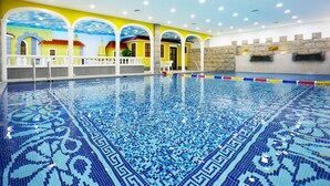 Une piscine intérieure dotée d’un sol en mosaïque bleue, de rampes blanches et d’une fresque jaune et bleue sur les murs.
