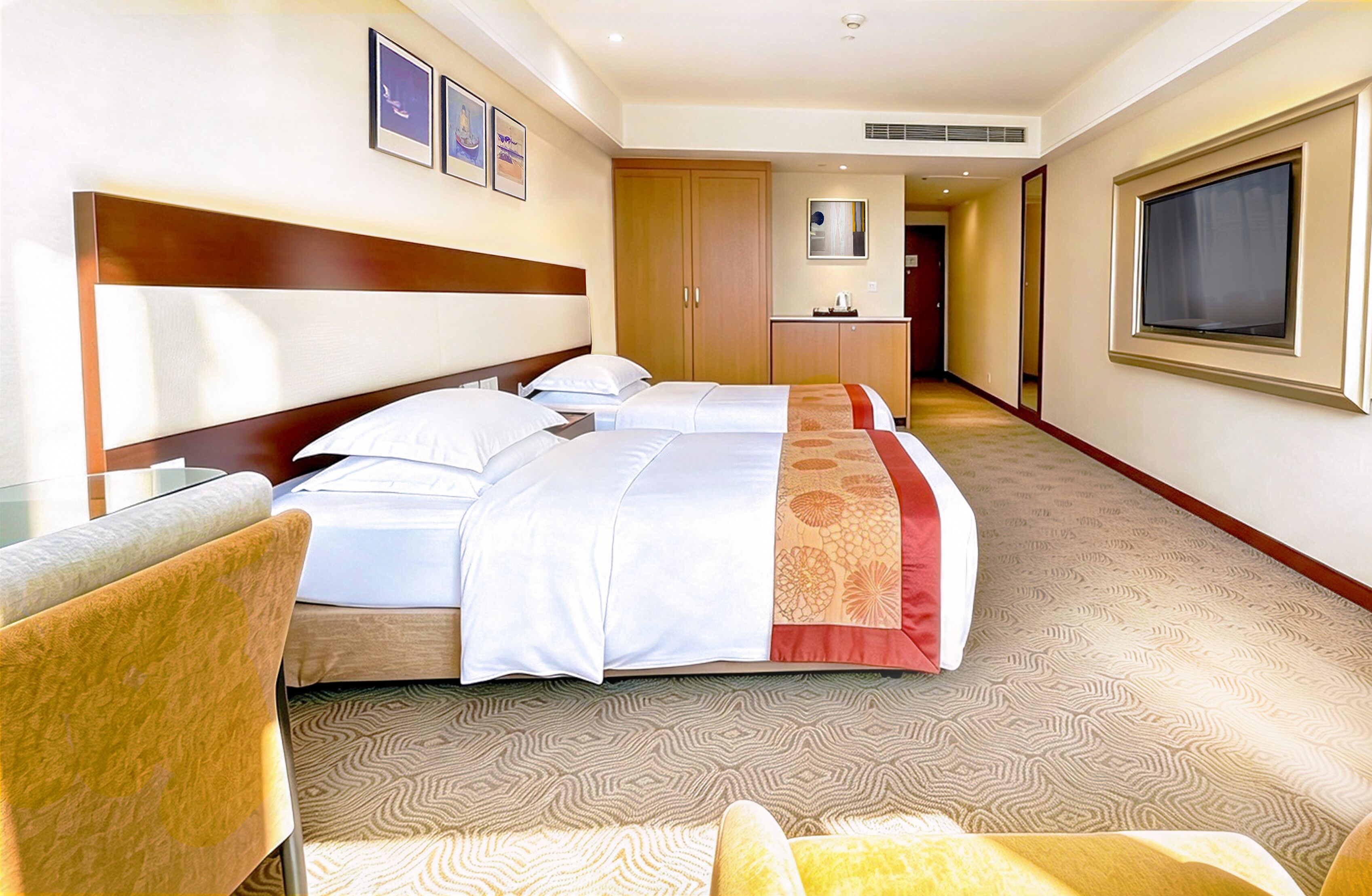 A hotel room with a large bed, a desk, and a chair. There are framed pictures on the wall and a window on the right.