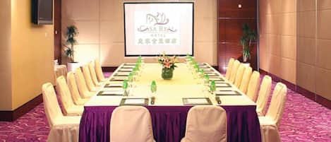 Un salón de conferencias con una mesa larga, sillas y una pantalla de proyector mostrando 'Casa Real Hotel'.