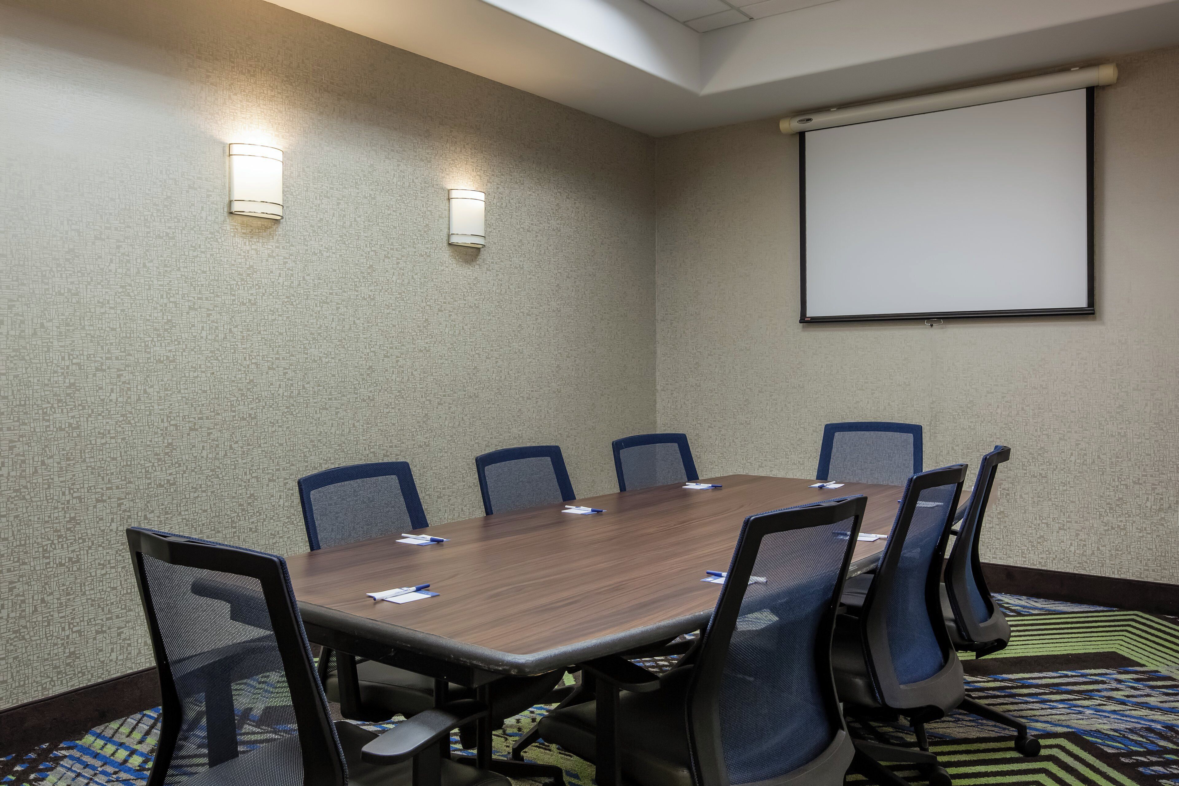 A conference room with a long table, six chairs, a projector screen, and two wall-mounted lights.