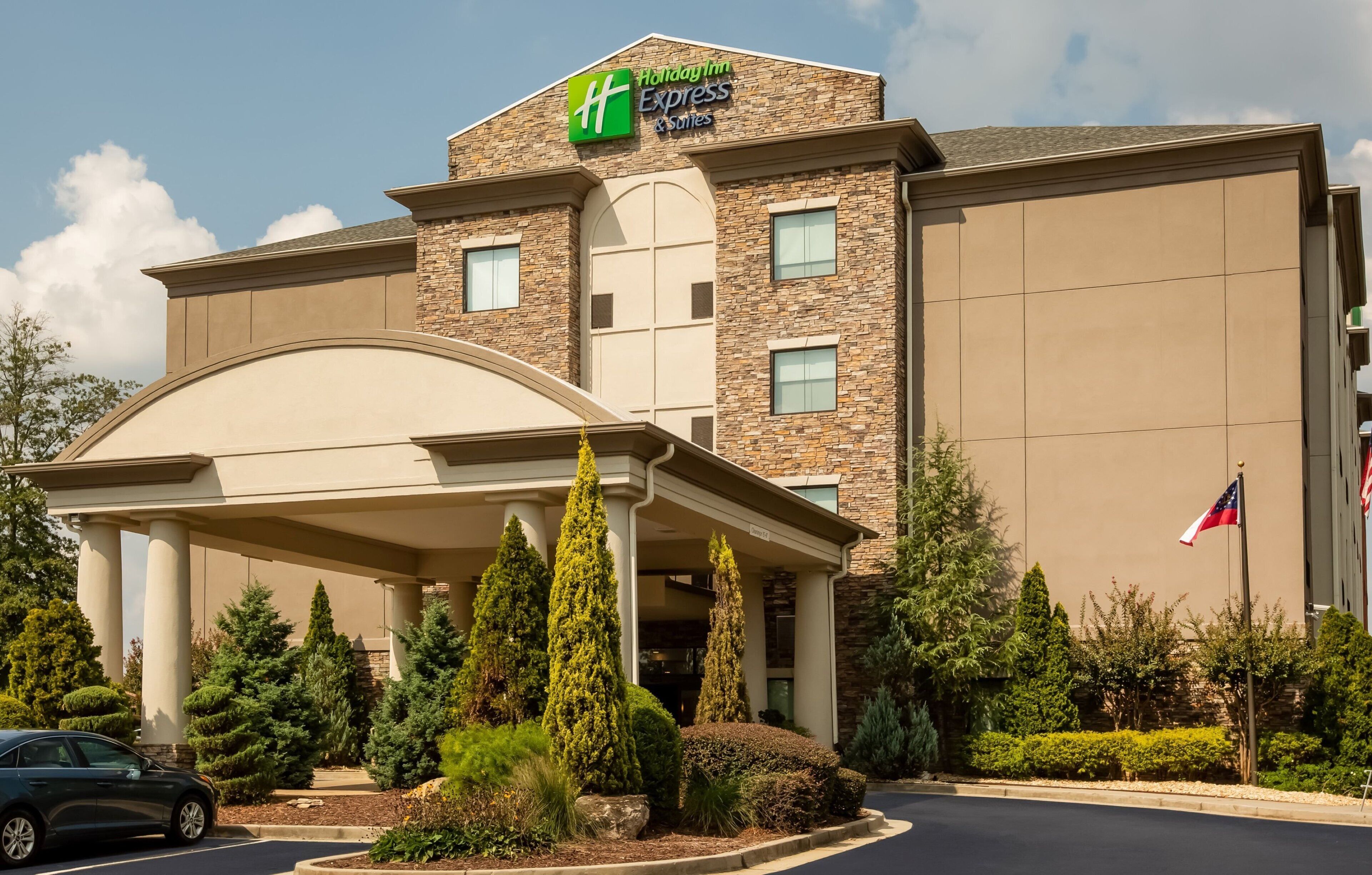 Edificio de Holiday Inn Express & Suites con entrada cubierta, fachada de piedra y bandera estadounidense.