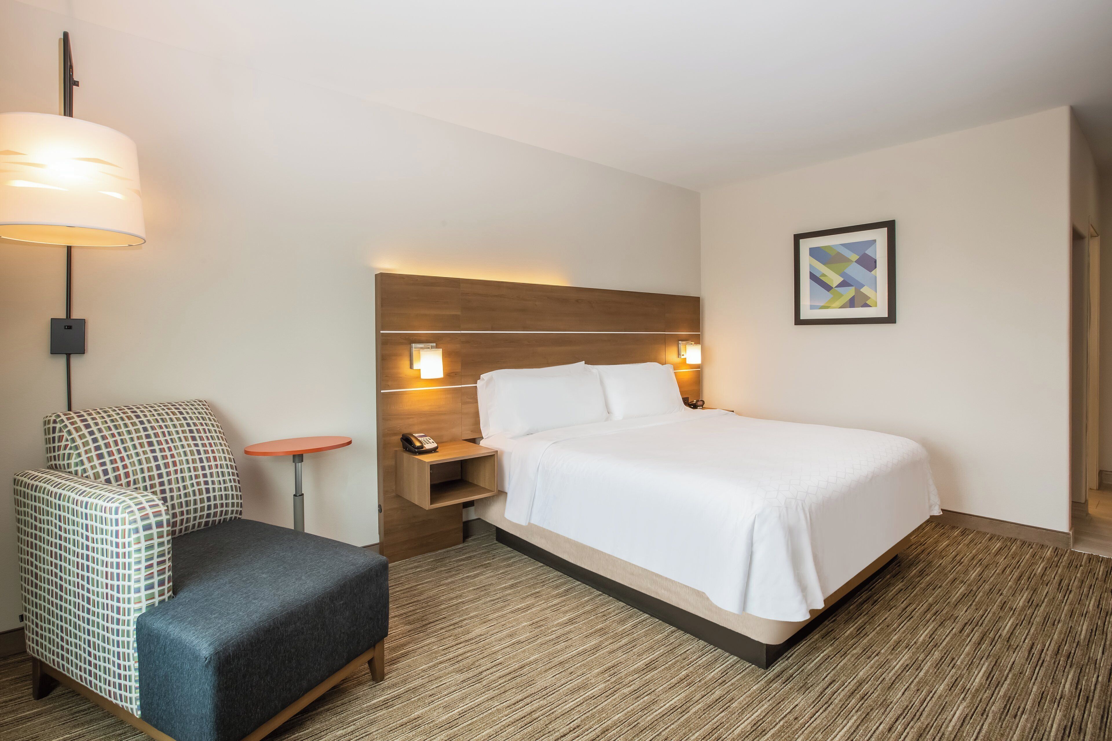 A hotel room with a bed, a chair, a bedside table with a lamp, and a framed picture on the wall.