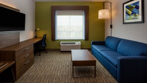 A modern hotel room with a wooden desk, a blue sofa, a flat-screen TV mounted on the wall, a large window with a view, and a framed abstract painting. - Holiday Inn Express Hotel & Suites Fairburn by IHG (Fairburn)
