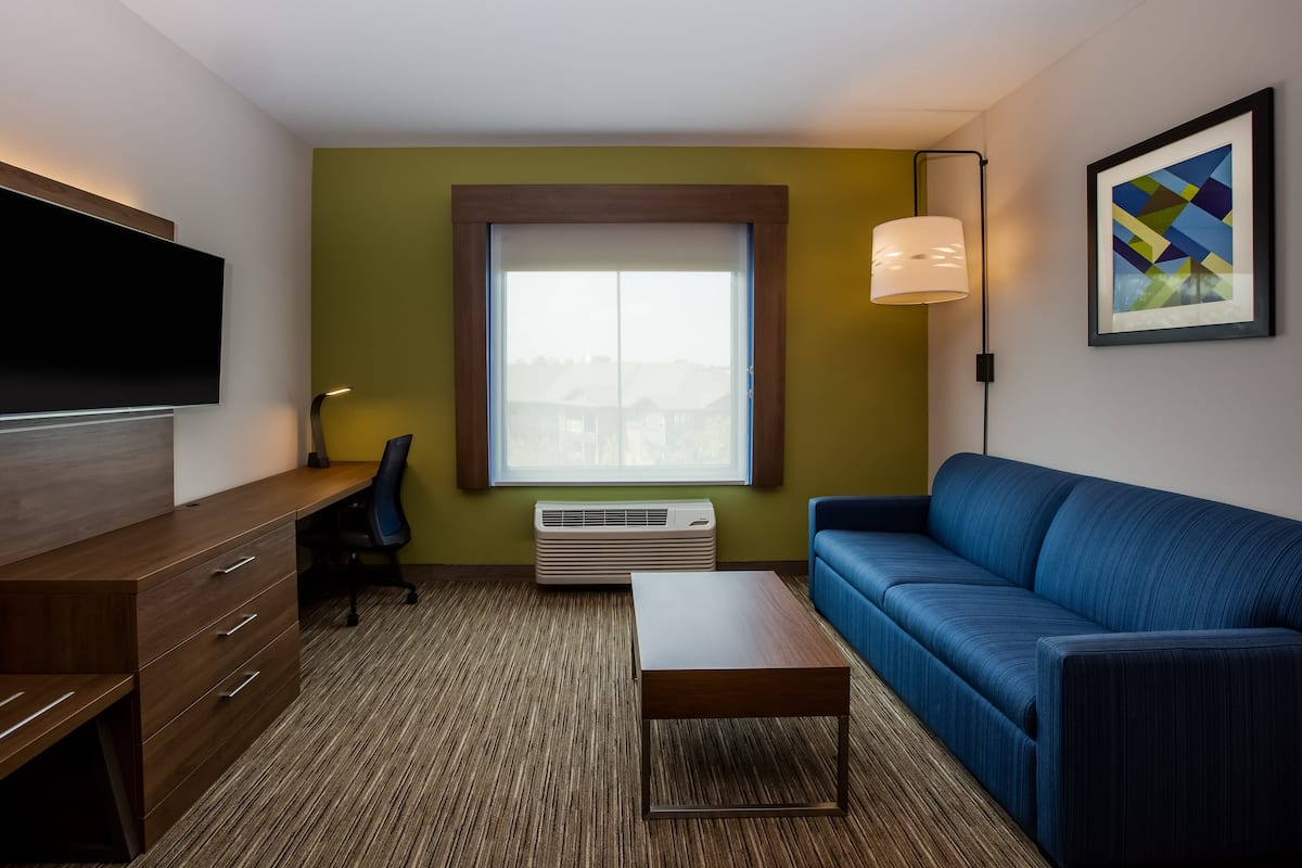 A modern hotel room with a wooden desk, a blue sofa, a flat-screen TV mounted on the wall, a large window with a view, and a framed abstract painting.
