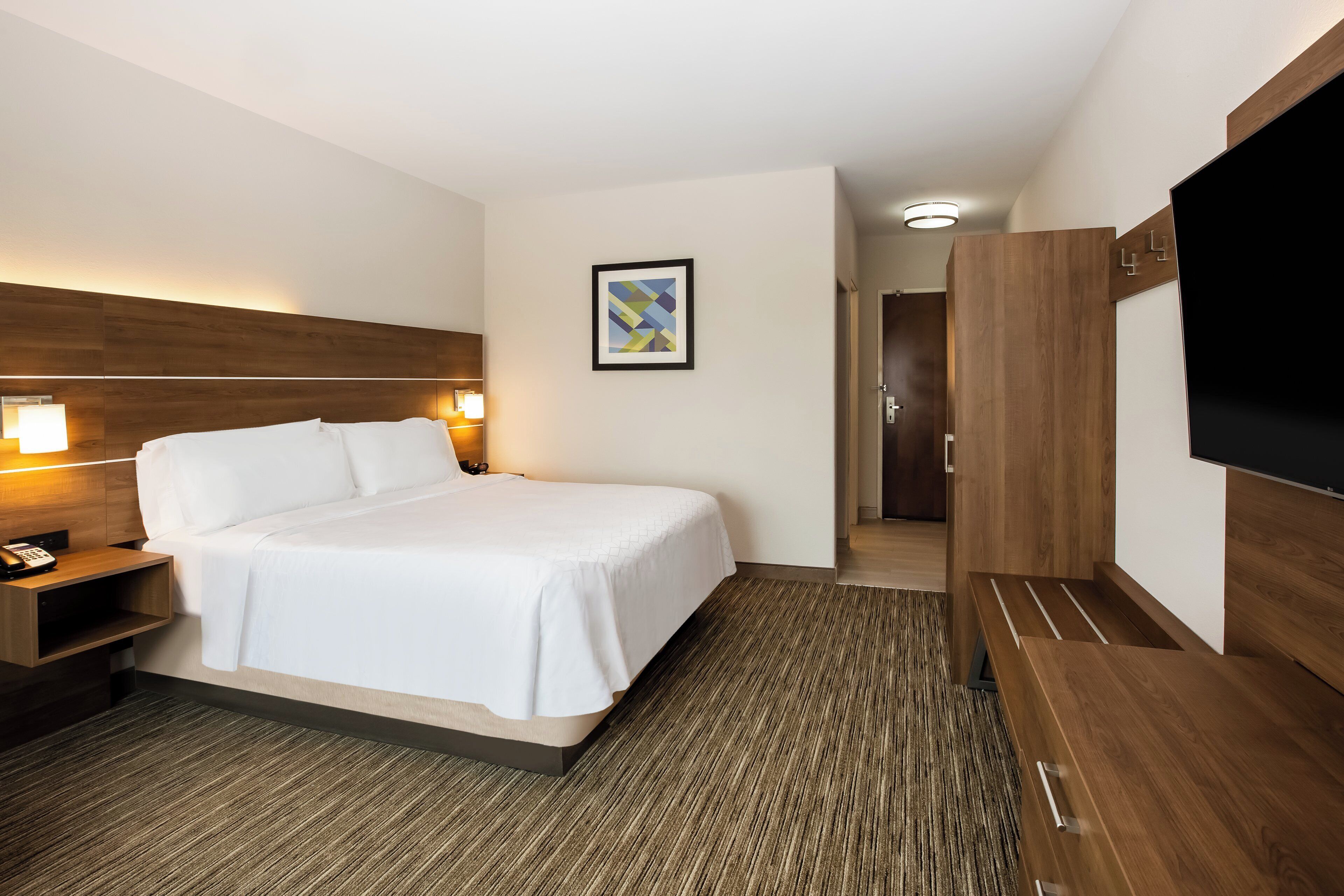 Une chambre d’hôtel avec un lit, des lampes de chevet, un bureau avec un téléphone, une télévision et un tableau encadré au mur.