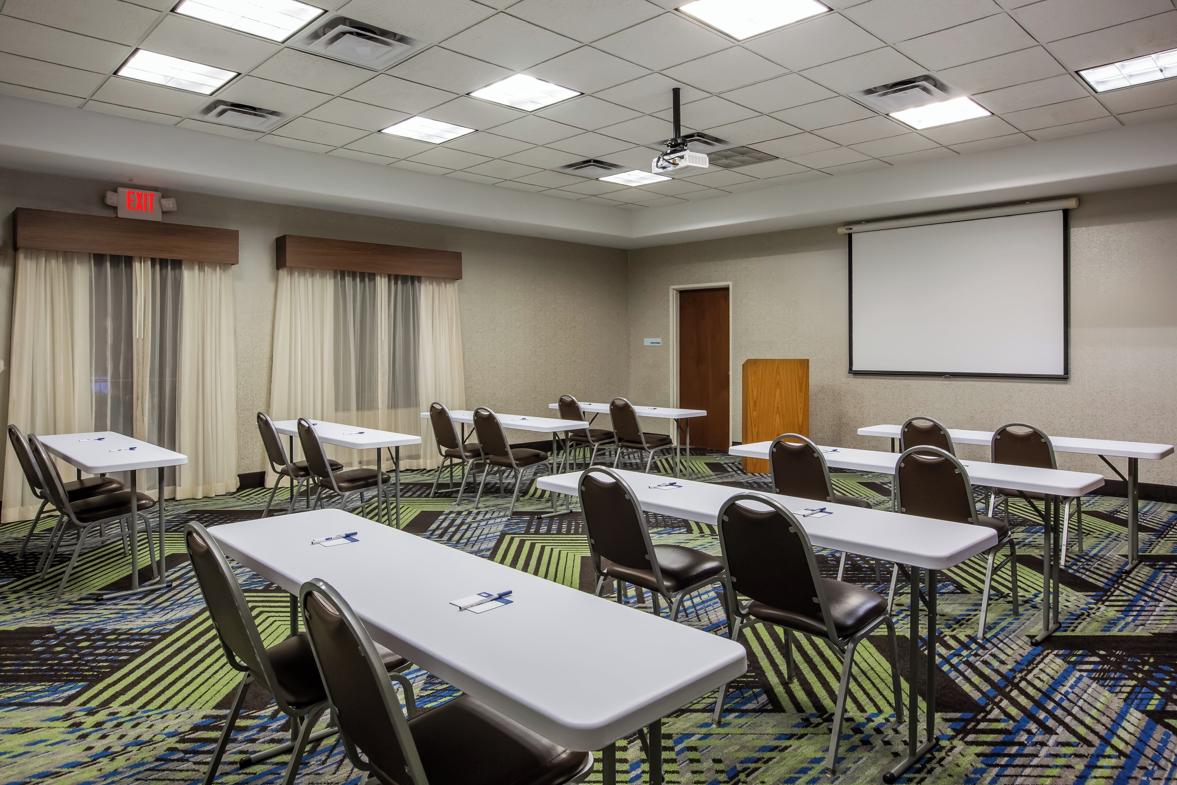 A conference room with multiple tables and chairs, a projector screen, and a carpeted floor.