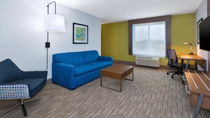 Quarto de hotel moderno com sofá azul, mesa de centro de madeira, escrivaninha com cadeira, televisão e uma janela grande.