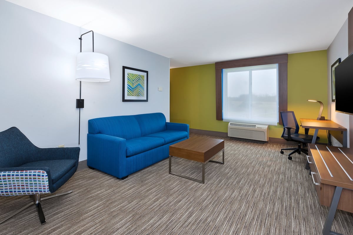 Een moderne hotelkamer met een blauwe bank, een houten salontafel, een bureau met stoel, een televisie en een groot raam.