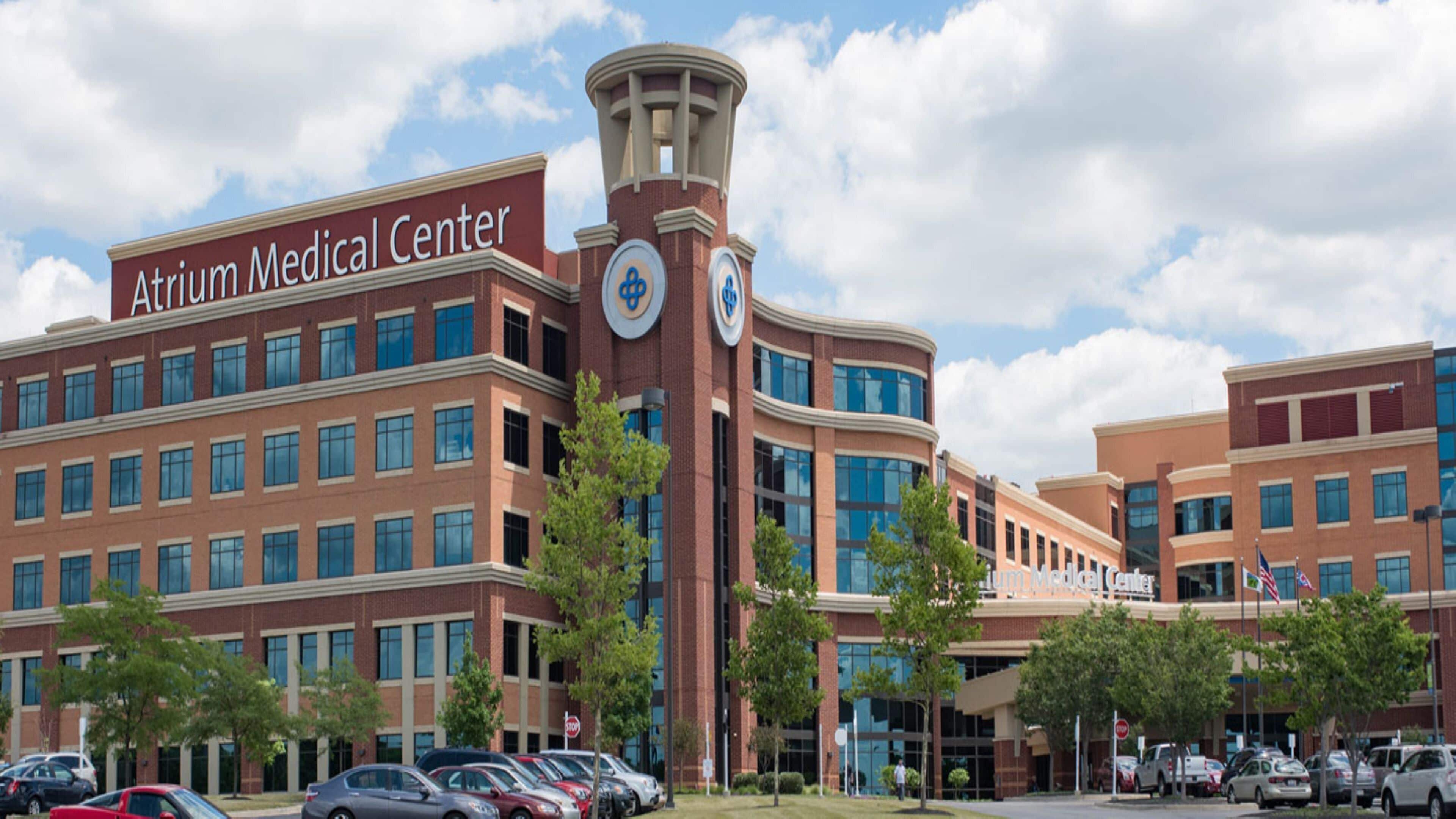 Un edificio pluripiano dell'Atrium Medical Center con una torre dell'orologio ben visibile, un parcheggio e cielo sereno.