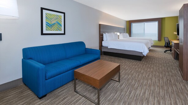 Quarto de hotel com sofá azul, mesa de centro de madeira, cama com roupa de cama branca, uma janela grande e uma televisão.