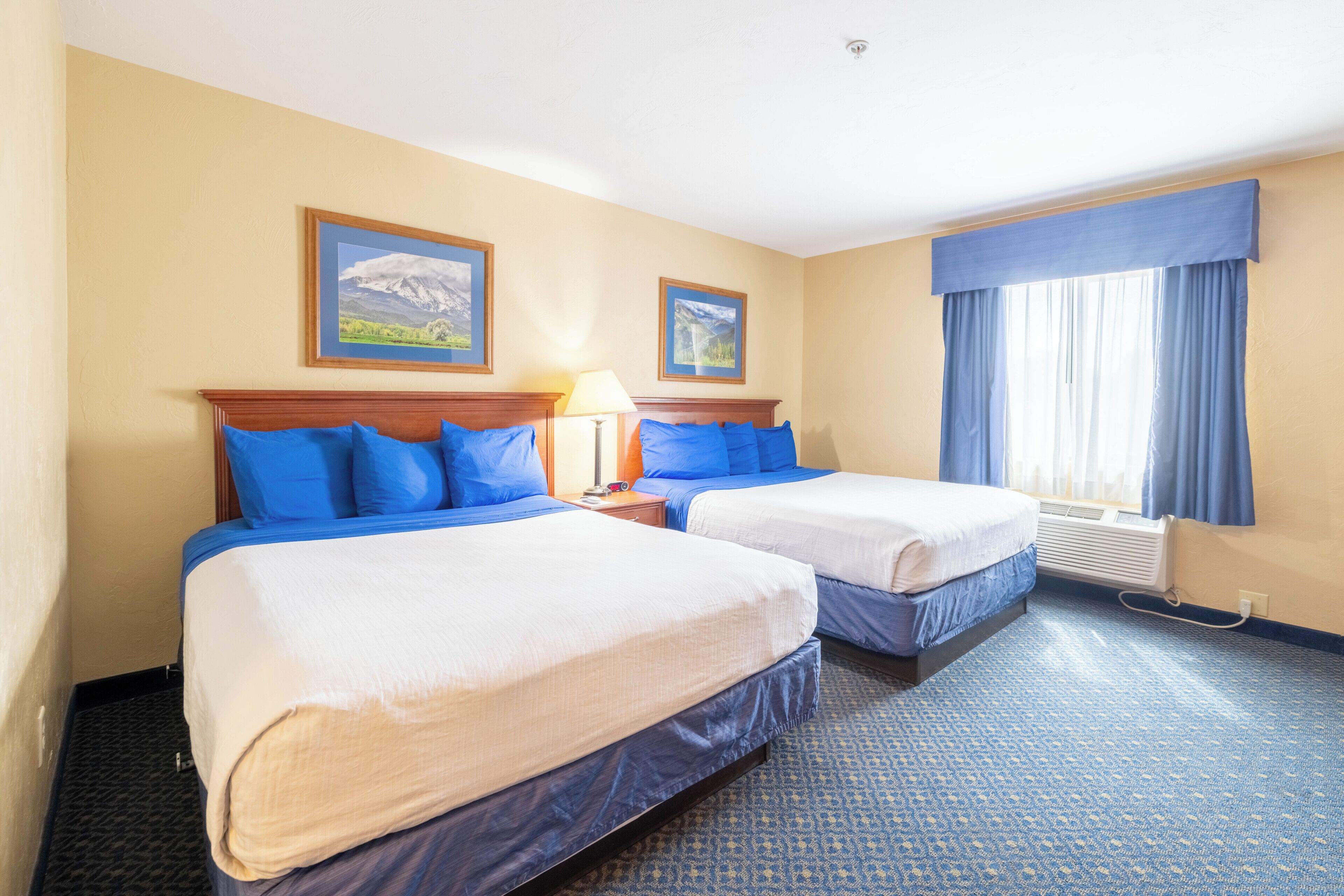 Una habitación de hotel con dos camas, una mesita de noche, una lámpara, una ventana con cortinas y un calentador fijado a la pared.
