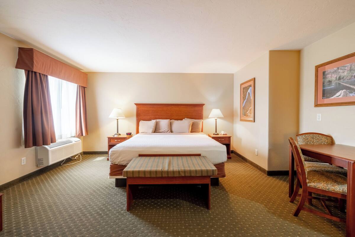 A hotel room with a large bed, bedside lamps, a desk with chairs, and a window with curtains.
