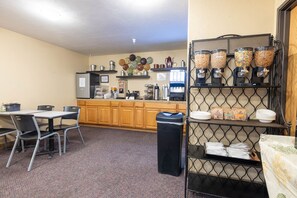 A small kitchenette with a coffee machine, a microwave, and a refrigerator. There is a table with chairs and a trash bin.