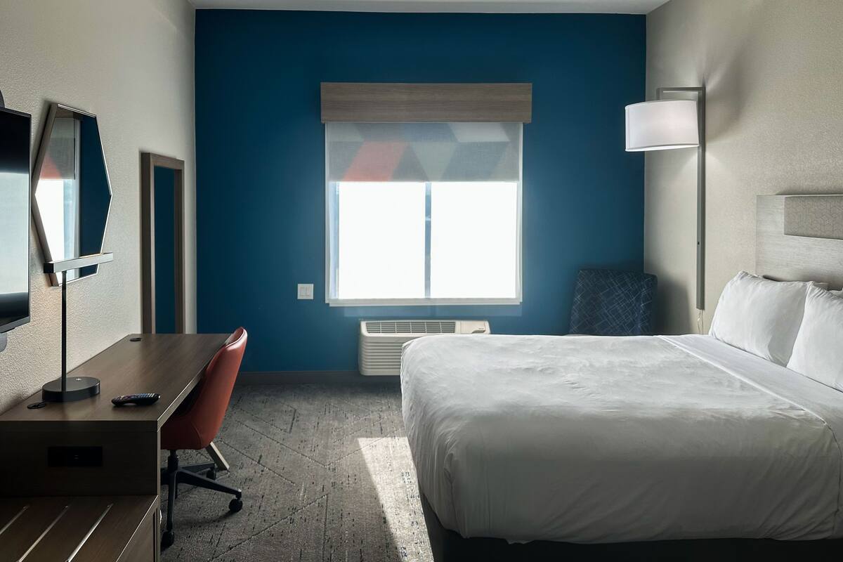 Ett hotellrum med en säng, ett skrivbord, en stol, en TV och ett fönster med persienner.