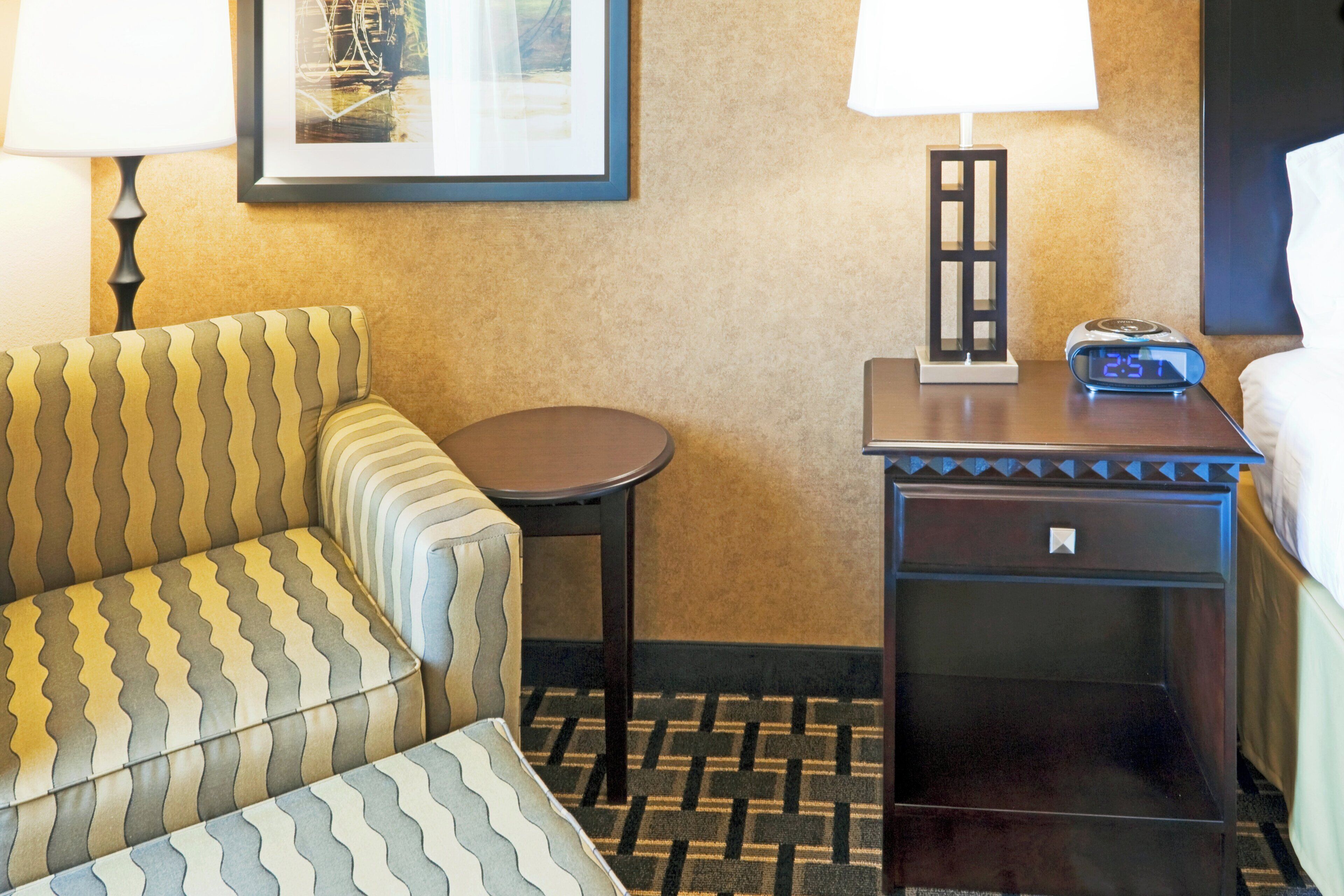 Une chambre d’hôtel avec un fauteuil à rayures, une petite table ronde, une table de chevet dotée d’une horloge numérique et un tableau encadré accroché au mur.