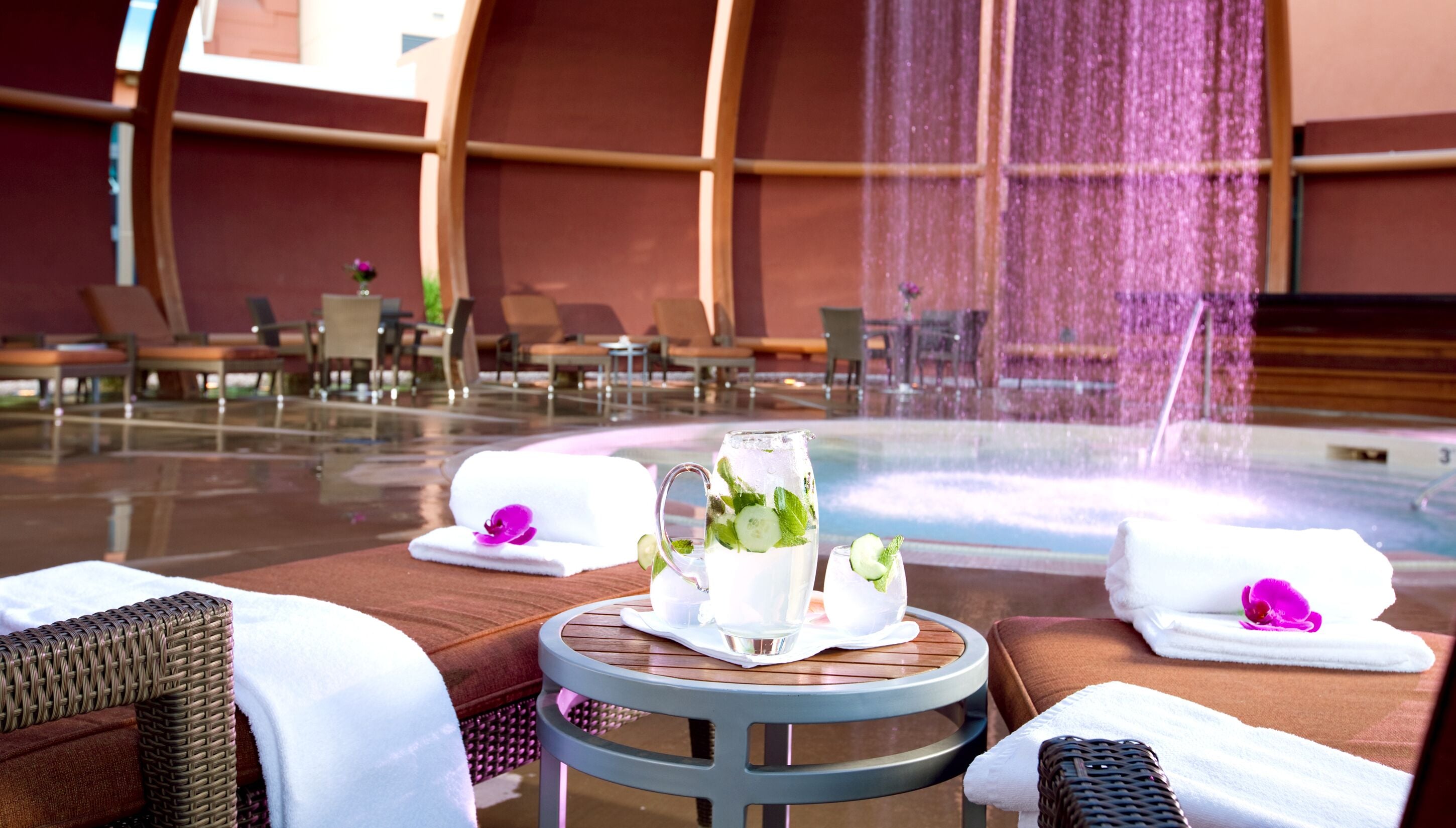 Área junto à piscina com espreguiçadeiras, uma mesa pequena com jarra de água e folhas de hortelã, e um elemento decorativo com água ao fundo.
