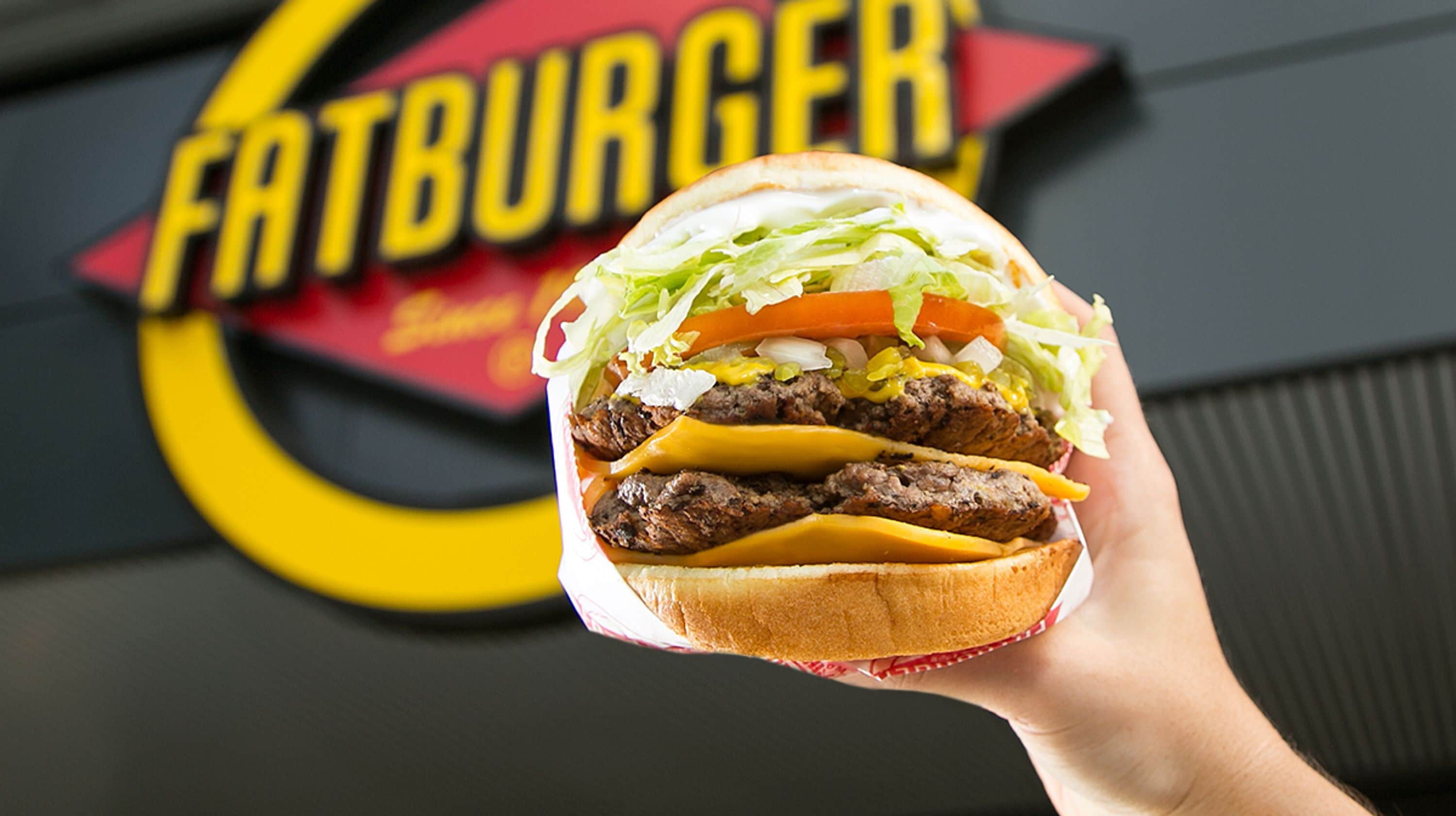 A hand holding a burger with visible lettuce, tomato, and cheese, in front of a 'Fatburger' sign.