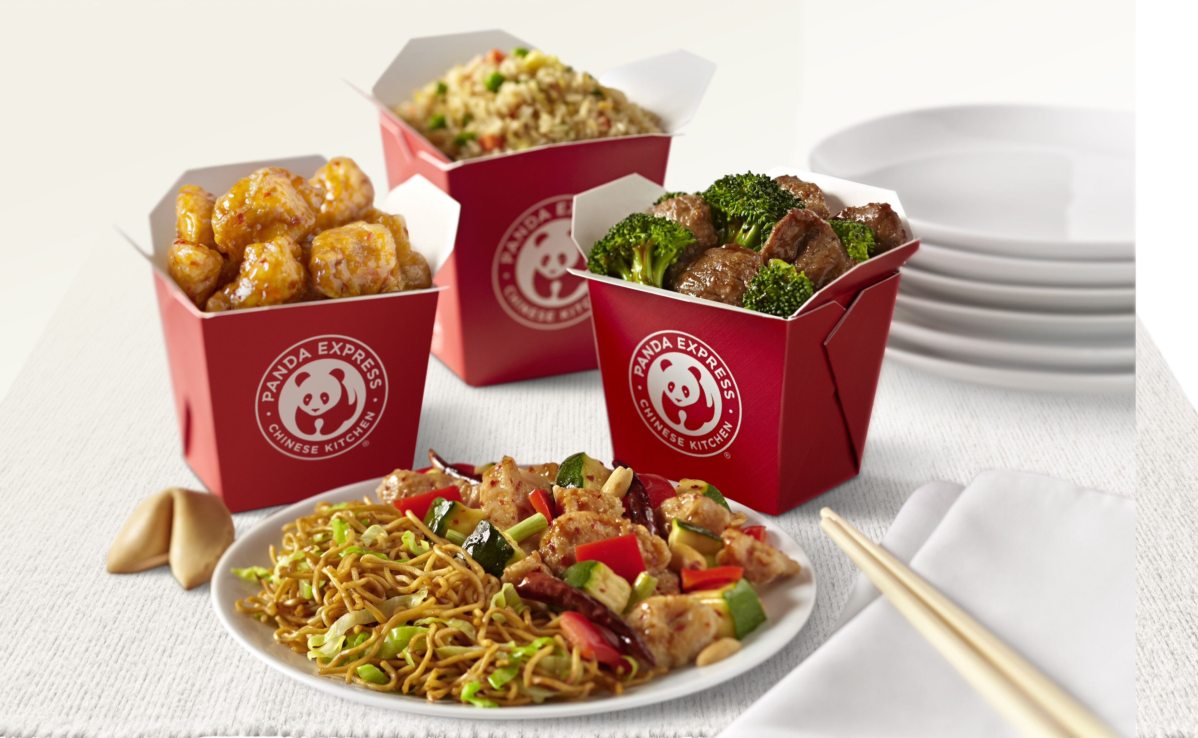 Une table avec quatre boîtes de nourriture de Panda Express, une assiette de nouilles sautées et une paire de baguettes.