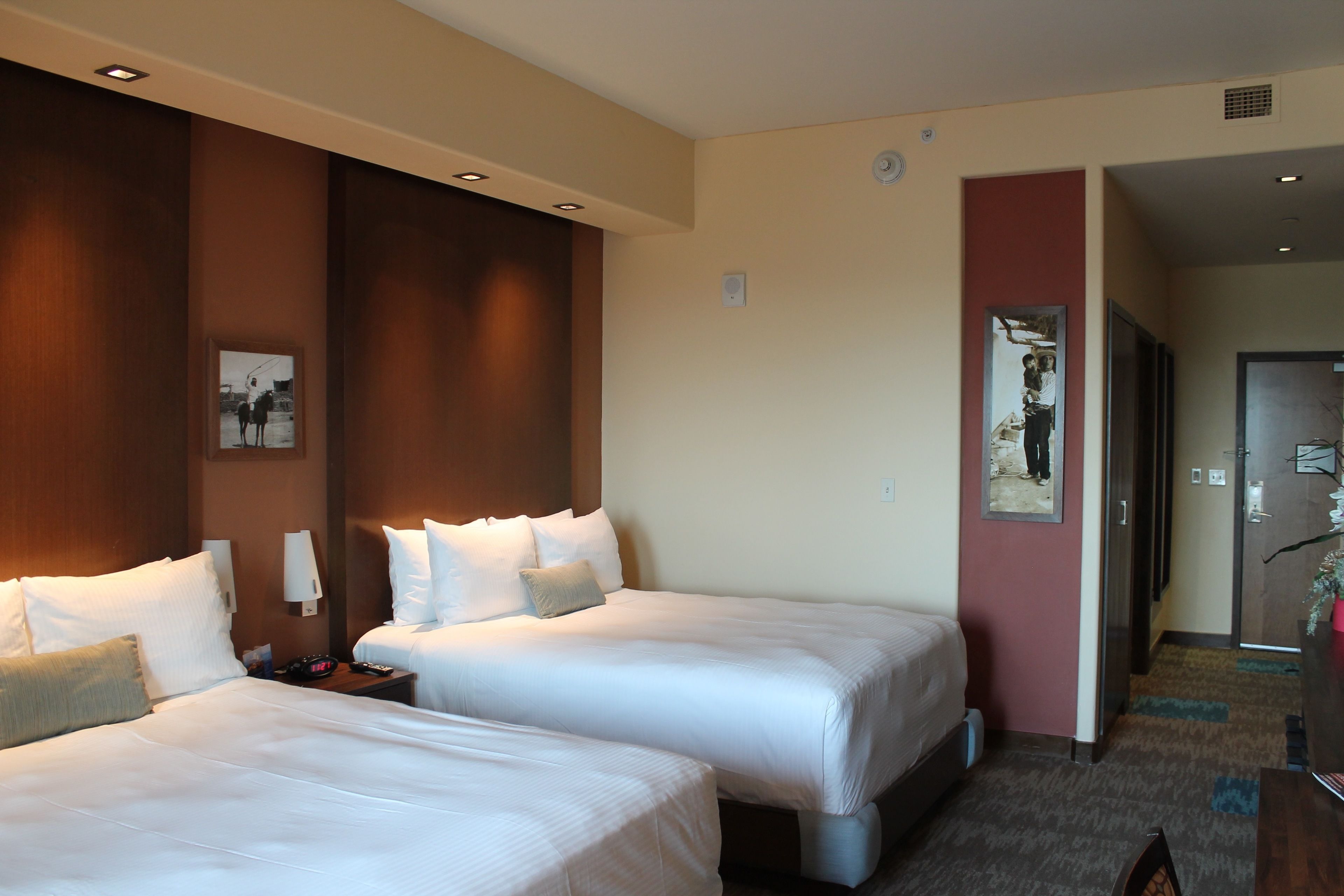 A hotel room with two beds, a nightstand, a wall-mounted lamp, and a door leading to another room.