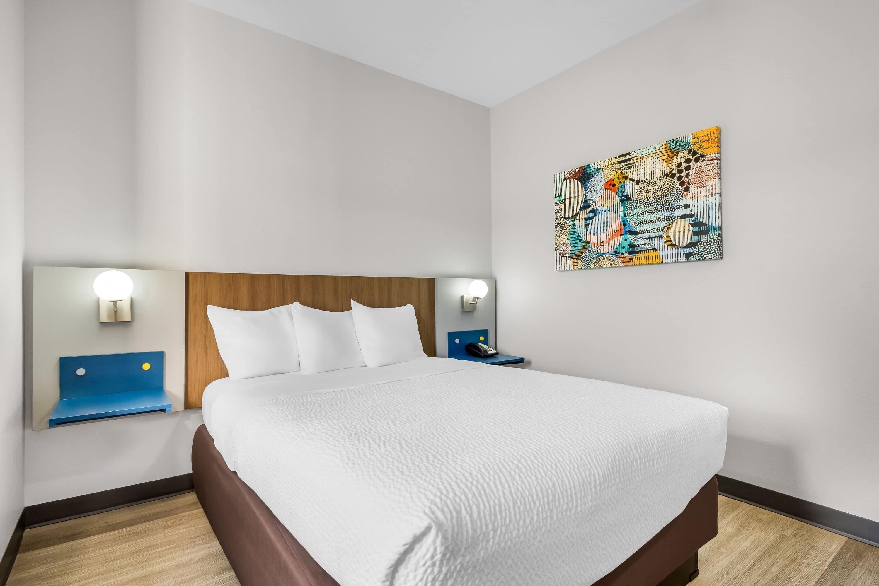Una habitación de hotel con una cama grande, dos mesitas de noche, un cabecero, una obra de arte colgada en la pared y un velador con lámpara.