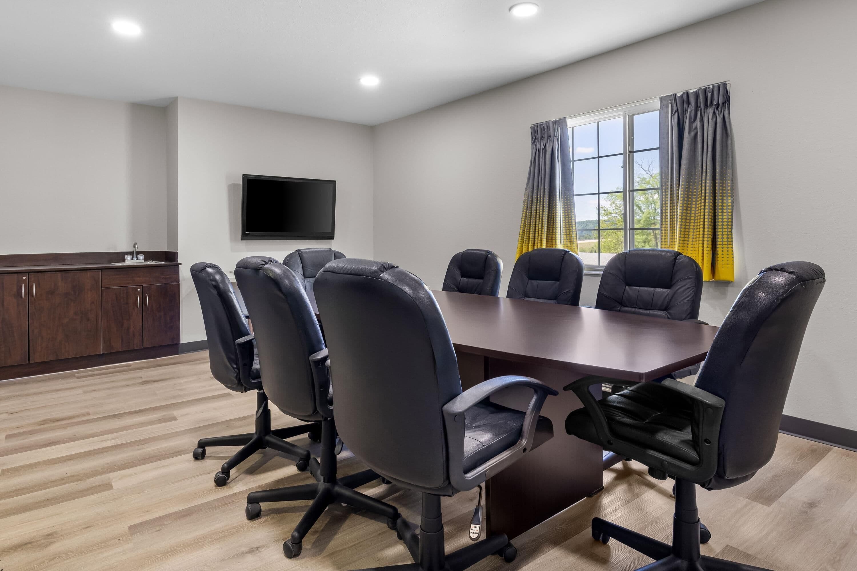 A conference room with a long table, six black office chairs, a flat-screen TV on the wall, and a window with curtains.